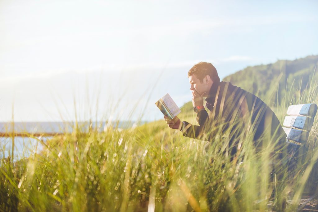 Homem sentado em um banco lendo um livro. Aparentemente, ele está sentado de frente ao mar, em um dia ensolarado com uma vegetação rasteira ao seu redor.