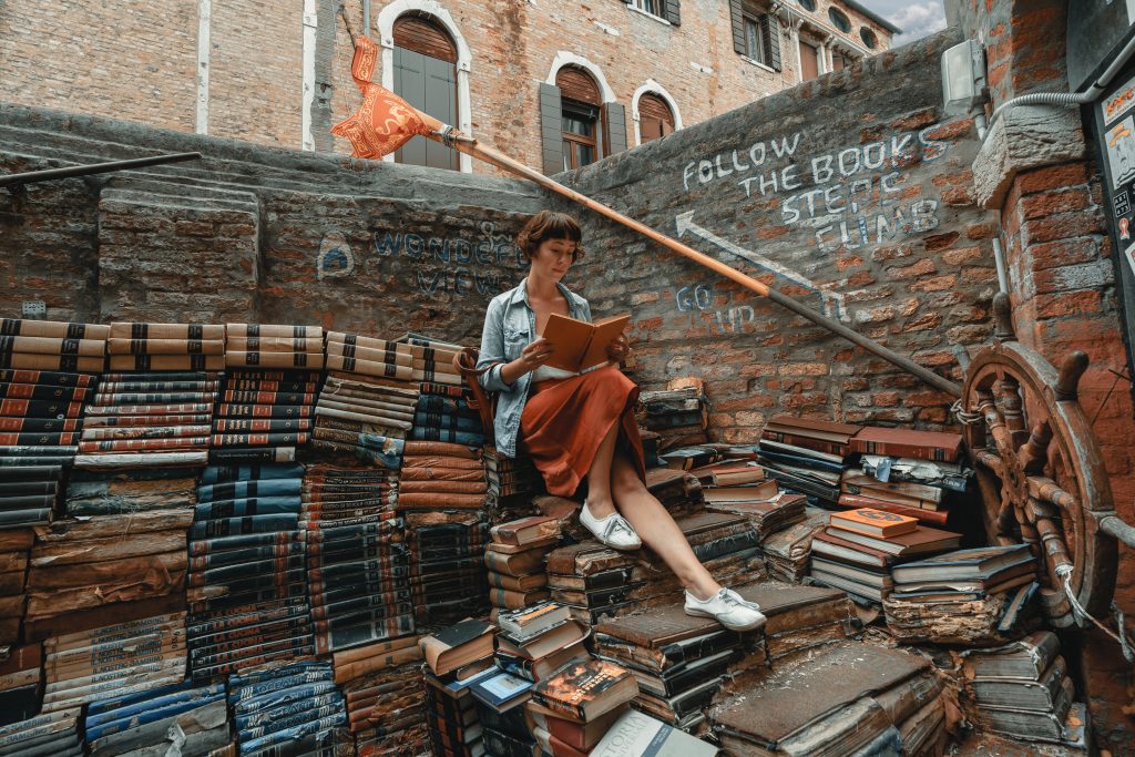 Mulher lendo sentada em uma pilha de livros na rua (quem sabe pensando em publicar um livro?).