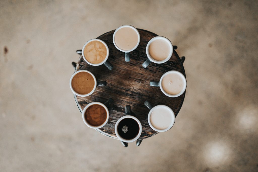 Na imagem, vemos uma diversidade de cafés, com cores diferentes em canecas, vistos de cima sobre uma mesa.