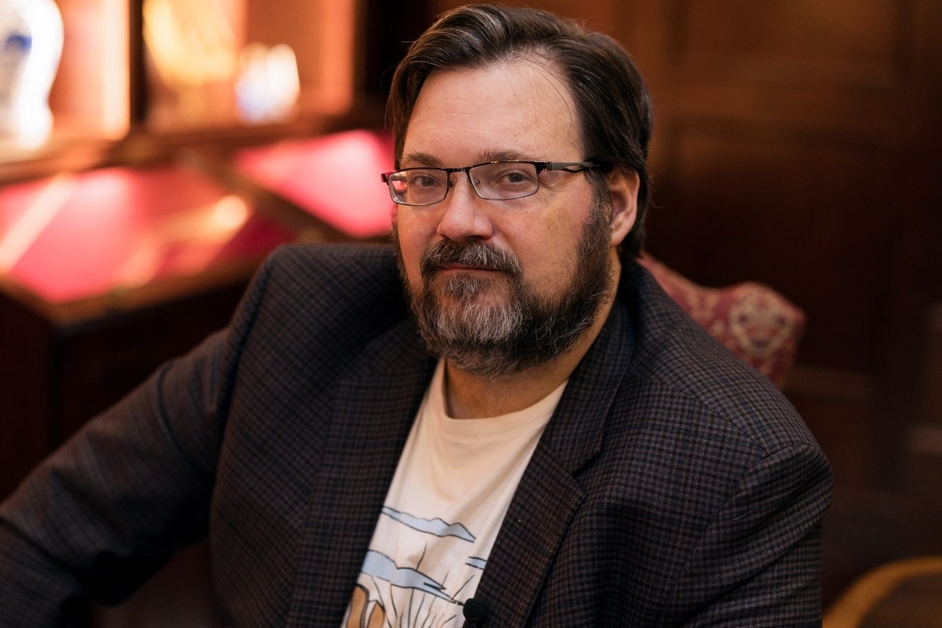 Na foto, o autor Brandon Sanderson está sentado em um ambiente elegante, provavelmente uma sala com móveis de madeira escura e iluminação quente. Ele veste um blazer em tons escuros sobre uma camiseta com uma estampa de montanhas e um sol. Sanderson tem cabelos castanhos escuros, um pouco longos, e uma barba cheia. Seus óculos de armação fina descansam sobre seu nariz, e ele olha diretamente para a câmera com uma expressão serena. Ao fundo, há prateleiras com objetos decorativos, iluminados suavemente, o que confere à cena um tom acolhedor e sofisticado.